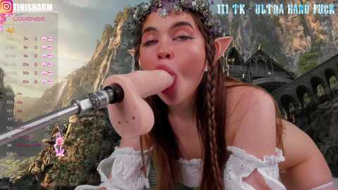 Media: Video of a fair-skinned, brown-haired woman with pointed elf ears, wearing a white off-shoulder dress and floral crown, performing oral sex on a large, realistic dildo. Background features a lush, fantasy landscape with a castle.