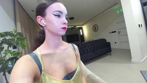 Media: Video of a slim, fair-skinned woman with dark hair in a ponytail, wearing a yellow tank top and black bra, standing in a modern living room with a dark couch, plants, and abstract art on the wall.