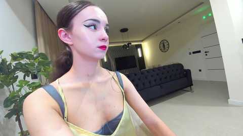 Media: Video of a fair-skinned woman with brown hair in a ponytail, wearing a yellow spaghetti-strap top, standing in a modern living room with a black sofa, green plant, and minimalist decor.