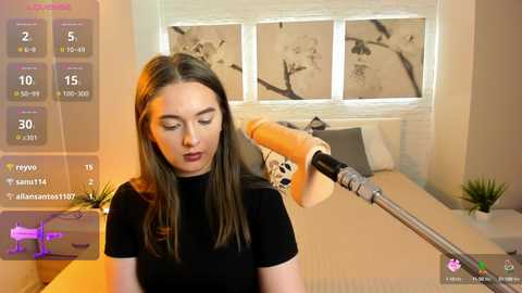 Media: Video of a young Caucasian woman with long brown hair, wearing a black top, holding a dildo-shaped vibrator in a modern bedroom with minimalist decor.