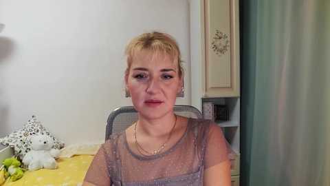 Media: Video of a young woman with short blonde hair, wearing a sheer, polka-dotted top, seated indoors against a white wall with floral patterns.