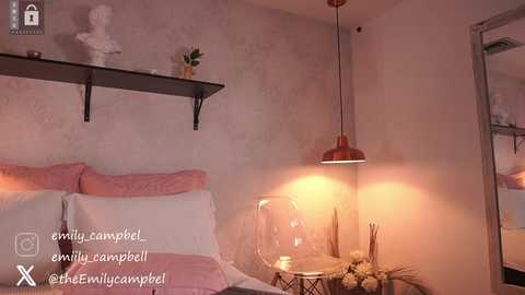 Media: Video of a cozy bedroom with a pink bed, white pillows, a wall-mounted shelf, a black metal lamp, and a framed mirror. The room features soft, warm lighting and a subtle, textured wallpaper.