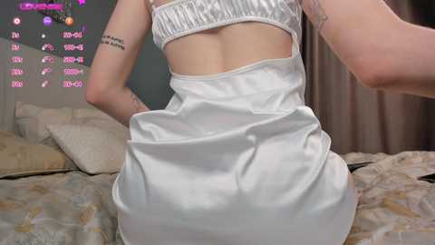 Media: Video of a slender, fair-skinned woman in a white satin mini-dress, showcasing her backside and revealing a tattoo on her left arm. Background features a bed with beige bedding and a purple light display.