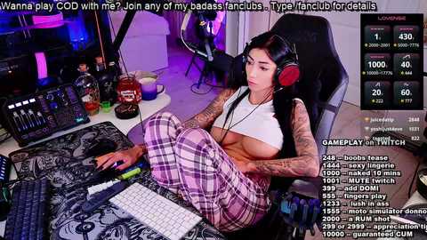 Media: Video of a tattooed, partially nude woman in plaid pajamas, seated at a gaming desk. Background includes gaming equipment and Twitch chat logs.