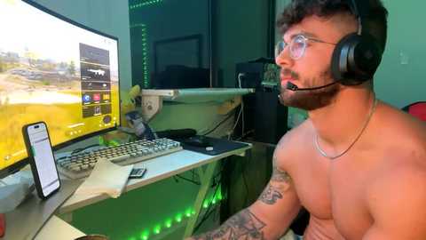 Media: Video of a shirtless, tattooed man with glasses and headphones, playing video games on a green-lit desk in a dimly lit room.