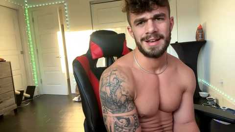 Media: Video of a shirtless, muscular man with a tattooed arm and short, curly hair, sitting in a black gaming chair with red accents, in a modern, dimly lit room with green LED strip lights and a desk cluttered with gaming accessories.