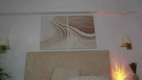 Media: Video of a minimalist bedroom featuring a beige upholstered headboard, beige bedding, and a large abstract wall art with wavy lines. Two modern wall sconces with beige shades flank the art, and a small, glowing candle on the nightstand adds a soft ambiance.