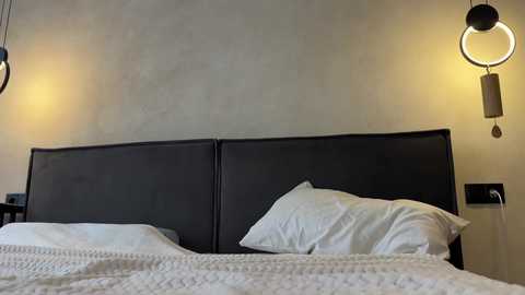 Media: Video of a modern, minimalist bedroom with a dark gray upholstered headboard, white bedding, and two pendant lights with circular shades on either side.