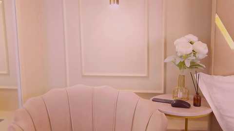 Media: Video of a chic, minimalist bedroom featuring a soft pink shell chair, a small round table with a vase of white flowers, and a beige wall with a framed window.