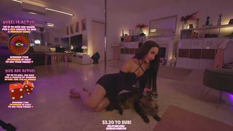 Media: Video of a woman with long black hair and dark skin, wearing a black off-shoulder dress, kneeling on a carpeted floor in a modern, dimly-lit living room with cats.