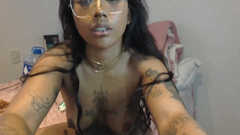 Media: A video of a young, dark-skinned woman with long, curly hair, wearing glasses, and showing tattoos on her arms and chest, standing in a bedroom.