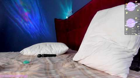 Media: A video of a neatly made bed with white pillows and a black vibrator on a plaid blanket, against a blue and red wall with glowing aurora lights, in a cozy, intimate bedroom setting.