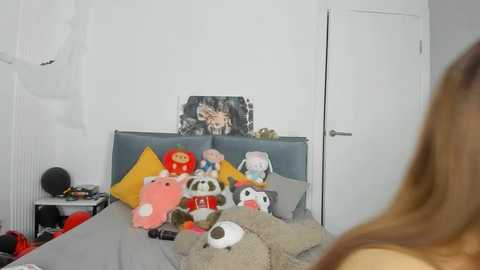 Media: Video of a cluttered bedroom with a woman's back visible. Bed features plush toys, including a panda, pig, and bear. White walls, door, and wall art with a black cat and red heart.