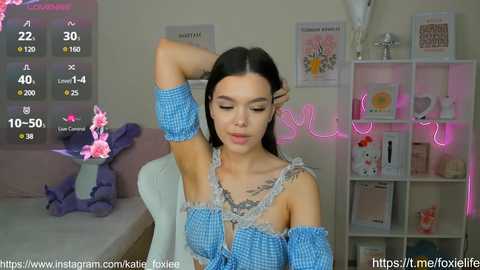 Media: Video of a young woman with fair skin, long dark hair, and a slender physique, wearing a blue checkered off-shoulder top, sitting in a cozy room adorned with pink neon lights and a plush toy.