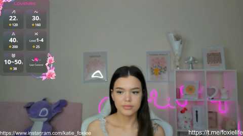 Media: Video of a young woman with long dark hair, sitting in a cozy, pink-themed room with a plush toy, a neon sign, and framed artwork.