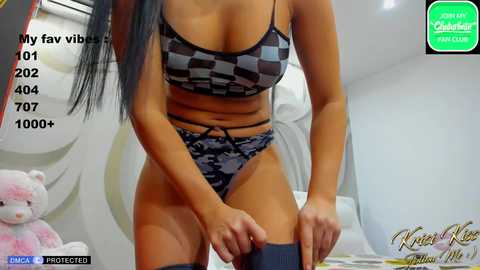 Media: A video of a busty, tan-skinned woman in a black and gray camo bra and matching panties, adjusting a dark blue sock, in a modern bedroom with white walls and a plush teddy bear.