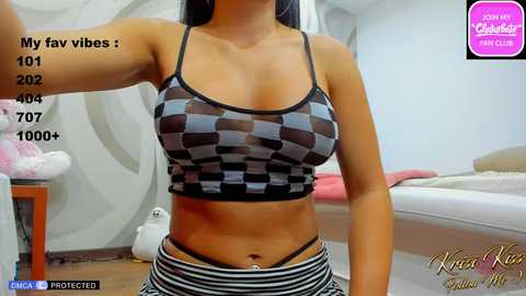 Media: Video of a woman with tan skin, long black hair, wearing a black and grey checkered crop top and striped shorts, standing in a modern bedroom with a white bed and white wall art.