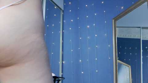 Media: A video of a partially visible person's bare upper chest and arm against a blue wall adorned with vertical, glowing string lights, framed mirrors reflecting the scene, and a visible ceiling.