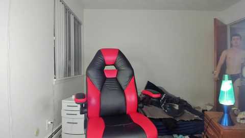 Media: Video of a modern gaming chair with red and black accents, positioned in a cluttered bedroom with a white dresser, open closet, and a man standing shirtless.