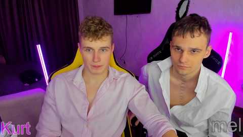 Media: Video of two young men with short hair, one with curly blonde hair and one with short brown hair, sitting in office chairs with purple neon lights behind them, wearing white shirts.