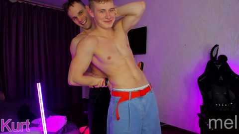 Media: Video of two shirtless, muscular men with fair skin and short hair, one wearing blue shorts, the other red belt, in a dimly lit room with purple and white lighting.