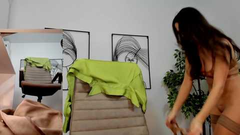 Media: Video of a nude woman with long dark hair adjusting a green towel draped over a beige chair in a minimalist room with black and white abstract art on the wall.