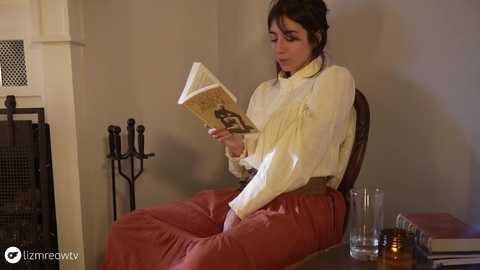 Media: Video of a young woman in a Victorian-style dress, reading a book in a dimly lit room with a fireplace, candle, and a glass of water.