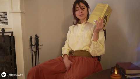 Media: Video of a young woman with long dark hair, wearing a cream blouse and red skirt, reading a book, seated in a cozy, dimly-lit room with a fireplace and books.