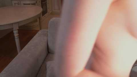 Media: Video of a nude person with light skin, sitting on a light gray couch in a room with a round white table and wooden floor.