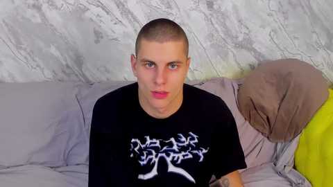 Media: Video of a bald, pale-skinned man with blue eyes, wearing a black T-shirt with white tree-like design, lying on a bed with grey sheets and a beige blanket, against a marble-patterned wall.