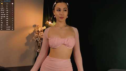 Media: Video of a young woman with light skin, dark hair, wearing a pastel pink, long-sleeved crop top and matching high-waisted pants, standing indoors against a dark background with a floral arrangement and digital clock.