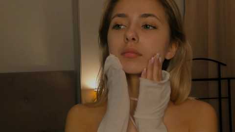 Media: Video of a young woman with light skin and blonde hair, wearing a white lace top, resting her chin on her hand, in a dimly lit room with wooden paneling and a lit lamp.