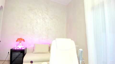 Media: Video of a minimalist living room with beige walls, a white armchair, a cream sofa, a black side table with a pink flower vase, and sheer white curtains.