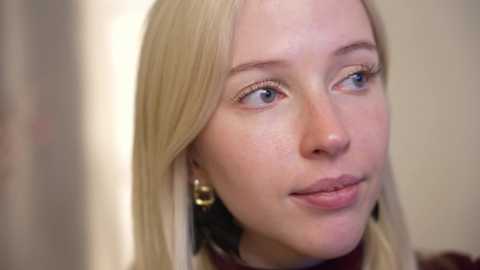 Media: A video of a fair-skinned woman with straight blonde hair, wearing subtle makeup, and black and gold hoop earrings. She gazes thoughtfully to the left, set against a blurred, neutral-colored background.