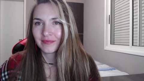 Media: A video of a young woman with long, straight, light brown hair, wearing a red and black plaid shirt, sitting in a room with a white window and blinds, and a bed visible in the background.