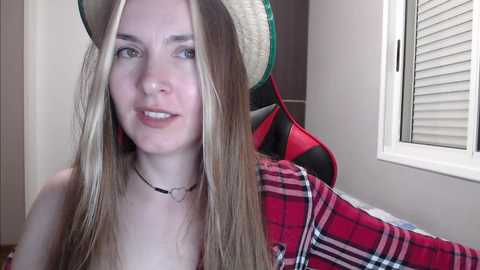 Media: A video of a young, light-skinned woman with long, straight blonde hair, wearing a straw hat and a red plaid shirt. She is smiling in a well-lit room with a window and a gaming chair in the background.