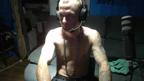 Media: Video of a shirtless, muscular man with a shaved head, wearing a headset, sitting in a dimly lit room with a green couch, a microphone stand, and a partially visible laptop.