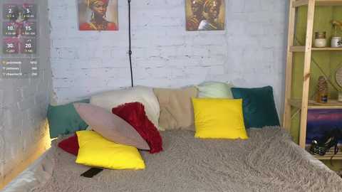 Media: Video of a cozy, bohemian bedroom with a white brick wall, vibrant cushions in red, yellow, and green, and a wooden shelf holding potted plants and a Buddha statue.