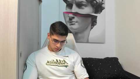 Media: A video of a young man with short dark hair and glasses, wearing a white shirt with a black graphic, sitting on a black cushioned chair. Behind him, a large black and white poster of Michelangelo's David.