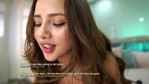 Media: Video of a young woman with light brown skin and long, wavy brown hair, wearing a light-colored top. She's smiling with closed eyes, set in a cozy bedroom with a bed and green walls.