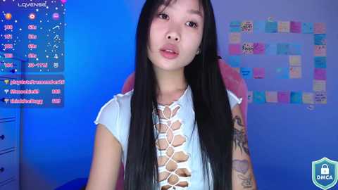 Media: Video of an Asian woman with long black hair, wearing a white lace-up top, sitting on a pink chair. Background features a blue wall with a colorful calendar and various notes.