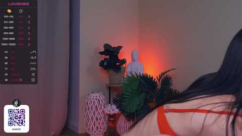 Media: A video showing a woman with long black hair, wearing a red bra, standing in a dimly lit room with a TV displaying a \"Live\" screen, featuring plants and candles.