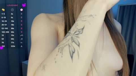 Media: Video of a topless woman with light skin, long brown hair, and a large butterfly tattoo on her right arm. She is indoors with blue curtains and a digital screen displaying social media metrics.