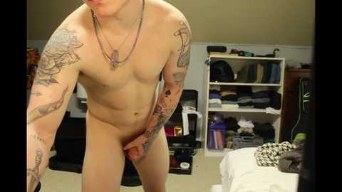 Media: A video of a nude, muscular, tattooed man with a circumcised penis, standing in a cluttered bedroom with beige walls, a white shelf, and a bed.