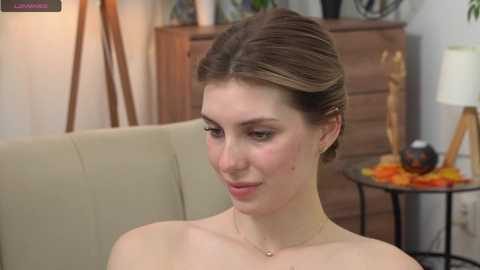 Media: Video of a young, fair-skinned woman with light brown hair in a neat bun, wearing a delicate necklace, sitting on a beige couch, in a cozy room with wooden furniture, a lamp, and plants in the background.