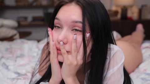 Media: Video of a young woman with long black hair, pale skin, and pink makeup, lying on a bed with a floral bedspread, resting her chin on her hands, looking sad.