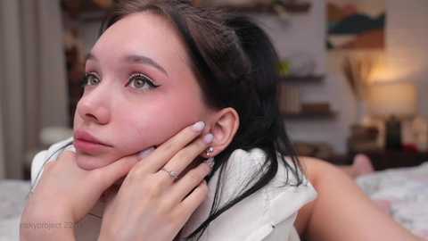 Media: Video of a young woman with fair skin, brown hair, and expressive eyes, lying on a bed, hand resting on her cheek, in a softly lit bedroom with a cozy atmosphere.