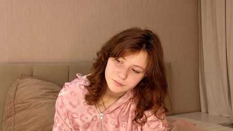 Media: A video of a young Caucasian woman with wavy brown hair, wearing a pink leopard print nightgown, sitting on a bed with beige pillows and a beige headboard, against a beige wall with vertical stripes.