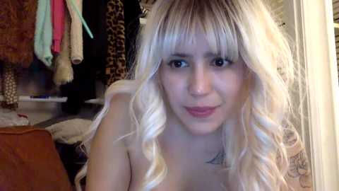 Media: Video of a young, blonde-haired woman with wavy hair, wearing a small, triangle-shaped tattoo on her chest. She has a neutral expression. Background shows a messy closet with leopard print and hanging clothes.