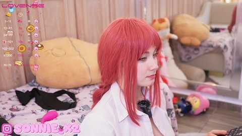 Media: Video of a young woman with red hair and fair skin in a plush, stuffed animal-filled room. She wears a white lab coat, sitting on a bed with a stuffed bear, surrounded by colorful toys.